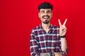 Young hispanic man with beard standing over red background smiling with happy face winking at the camera doing victory sign with Royalty Free Stock Photo