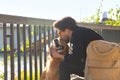 Young Hispanic man with beard, sunglasses, black shirt and backpack, sitting on a bench kissing his dog under the sun rays. Royalty Free Stock Photo