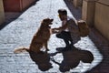 Young Hispanic man with beard and sunglasses crouching with his dog on a sunny street at sunset casting his shadow on the ground. Royalty Free Stock Photo