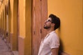 Young Hispanic man with beard and sunglasses leaning against a wall looking at the sky with distracted and thoughtful attitude. Royalty Free Stock Photo
