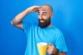 Young hispanic man with beard and tattoos drinking a cup of coffee very happy and smiling looking far away with hand over head Royalty Free Stock Photo