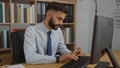 Young hispanic man with beard typing on computer keyboard in office workplace with bookshelves in the background Royalty Free Stock Photo