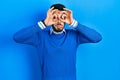 Young hispanic man with beard wearing casual blue sweater doing ok gesture like binoculars sticking tongue out, eyes looking Royalty Free Stock Photo