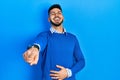 Young hispanic man with beard wearing casual blue sweater laughing at you, pointing finger to the camera with hand over body, Royalty Free Stock Photo