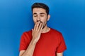 Young hispanic man with beard wearing red t shirt over blue background bored yawning tired covering mouth with hand Royalty Free Stock Photo