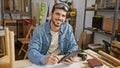 A young hispanic man with a beard wearing safety glasses and denim working with a tablet in a carpentry workshop Royalty Free Stock Photo