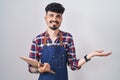 Young hispanic man with beard wearing waiter apron holding clipboard smiling cheerful presenting and pointing with palm of hand Royalty Free Stock Photo