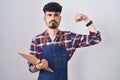 Young hispanic man with beard wearing waiter apron holding clipboard strong person showing arm muscle, confident and proud of