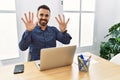 Young hispanic man with beard working at the office with laptop showing and pointing up with fingers number nine while smiling