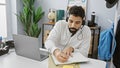 A young hispanic man with a beard working in an office, using a headset and writing notes on a notepad near his laptop Royalty Free Stock Photo