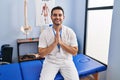 Young hispanic man with beard working at pain recovery clinic praying with hands together asking for forgiveness smiling confident Royalty Free Stock Photo