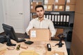 Young hispanic man with beard working at small business ecommerce holding box in shock face, looking skeptical and sarcastic, Royalty Free Stock Photo