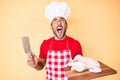 Young hispanic man cooking chicken holding knife angry and mad screaming frustrated and furious, shouting with anger looking up