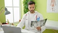 Young hispanic man doctor looking at xray typing on laptop at clinic