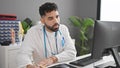 Young hispanic man doctor using computer writing notes at clinic Royalty Free Stock Photo