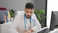 Young hispanic man doctor using computer writing notes at clinic Royalty Free Stock Photo