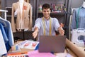 Young hispanic man dressmaker designer using laptop looking at the camera smiling with open arms for hug Royalty Free Stock Photo