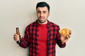 Young hispanic man drinking a bottle of beer and eating chip potatoes clueless and confused expression