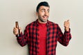 Young hispanic man drinking a bottle of beer pointing thumb up to the side smiling happy with open mouth Royalty Free Stock Photo