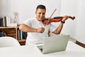 Young hispanic man having online violin lesson using laptop sitting on the table at home