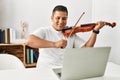 Young hispanic man having online violin lesson using laptop sitting on the table at home