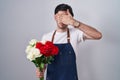 Young hispanic man holding bouquet of white and red roses covering eyes with hand, looking serious and sad Royalty Free Stock Photo