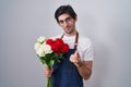 Young hispanic man holding bouquet of white and red roses doing money gesture with hands, asking for salary payment, millionaire