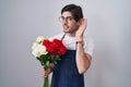 Young hispanic man holding bouquet of white and red roses smiling with hand over ear listening an hearing to rumor or gossip Royalty Free Stock Photo