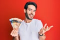 Young hispanic man holding canadian dollars celebrating achievement with happy smile and winner expression with raised hand