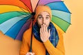 Young hispanic man holding colorful umbrella covering mouth with hand, shocked and afraid for mistake