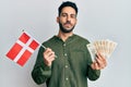 Young hispanic man holding denmark flag and krone banknotes puffing cheeks with funny face Royalty Free Stock Photo