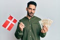 Young hispanic man holding denmark flag and krone banknotes relaxed with serious expression on face Royalty Free Stock Photo