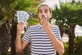 Young hispanic man holding dollars close to face covering mouth with hand, shocked and afraid for mistake Royalty Free Stock Photo