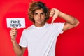 Young hispanic man holding fake news banner with angry face, negative sign showing dislike with thumbs down, rejection concept