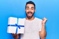 Young hispanic man holding gift screaming proud, celebrating victory and success very excited with raised arm