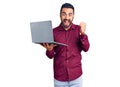 Young hispanic man holding laptop screaming proud, celebrating victory and success very excited with raised arms