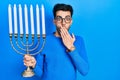 Young hispanic man holding menorah hanukkah jewish candle covering mouth with hand, shocked and afraid for mistake Royalty Free Stock Photo