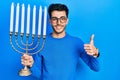 Young hispanic man holding menorah hanukkah jewish candle smiling happy and positive, thumb up doing excellent and approval sign Royalty Free Stock Photo