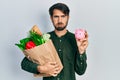 Young hispanic man holding piggy bank and paper bag with groceries puffing cheeks with funny face Royalty Free Stock Photo