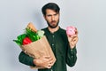 Young hispanic man holding piggy bank and paper bag with groceries in shock face, looking skeptical and sarcastic, surprised with Royalty Free Stock Photo