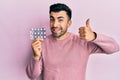 Young hispanic man holding pills smiling happy and positive, thumb up doing excellent and approval sign Royalty Free Stock Photo