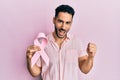 Young hispanic man holding pink cancer ribbon screaming proud, celebrating victory and success very excited with raised arms Royalty Free Stock Photo