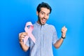 Young hispanic man holding pink cancer ribbon screaming proud, celebrating victory and success very excited with raised arms Royalty Free Stock Photo