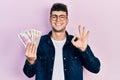 Young hispanic man holding saudi arabia riyal banknotes doing ok sign with fingers, smiling friendly gesturing excellent symbol Royalty Free Stock Photo