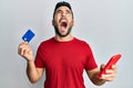 Young hispanic man holding smartphone and credit card angry and mad screaming frustrated and furious, shouting with anger looking Royalty Free Stock Photo