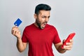 Young hispanic man holding smartphone and credit card angry and mad screaming frustrated and furious, shouting with anger Royalty Free Stock Photo