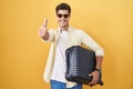 Young hispanic man holding suitcase going on summer vacation approving doing positive gesture with hand, thumbs up smiling and Royalty Free Stock Photo