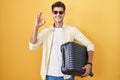 Young hispanic man holding suitcase going on summer vacation smiling positive doing ok sign with hand and fingers Royalty Free Stock Photo