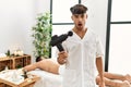 Young hispanic man holding therapy massage gun at wellness center scared and amazed with open mouth for surprise, disbelief face Royalty Free Stock Photo