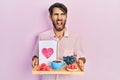 Young hispanic man holding tray with breakfast food and heart draw angry and mad screaming frustrated and furious, shouting with Royalty Free Stock Photo